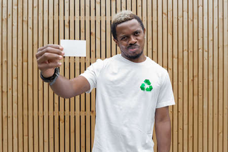 Young attractive man in t-shirt with recycle symbol on wooden background holding blank business card. Eco symbol. Minimalismの写真素材
