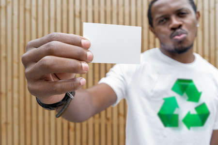 Young attractive man in t-shirt with recycle symbol on wooden background holding blank business card. Eco symbol. Minimalismの写真素材