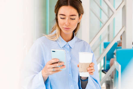 Young and attractive woman drinking coffee from coffee eco biodegradable cup at modern park and holding mobile phone. Eco friendly take away mug.の写真素材