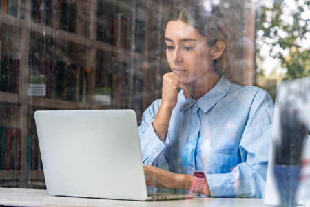 Young and pensive attractive woman working on laptop in modern library with big windows. Freelance concept. Young successful entrepreneur.の写真素材