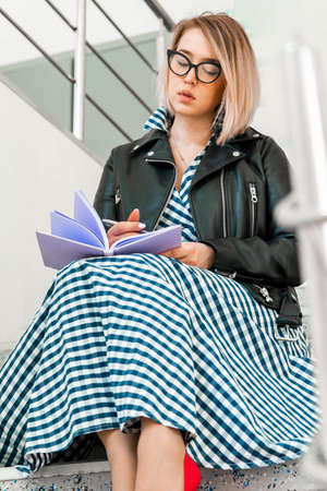 Young attractive blond woman makes some notes in violet notepad at library and sitting on ladder steps.の写真素材