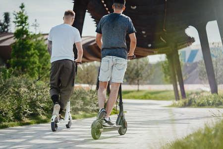Two men riding white and black electric kick scooters at beautiful park landscape. Men is on foreground, modern building and park is on background.の写真素材