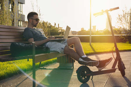Young attractive man with laptop and modern electric kick scooter sitting at contemporary park at bench. and working Freelancer or business man working on laptop. Beautiful Sunset time.の写真素材