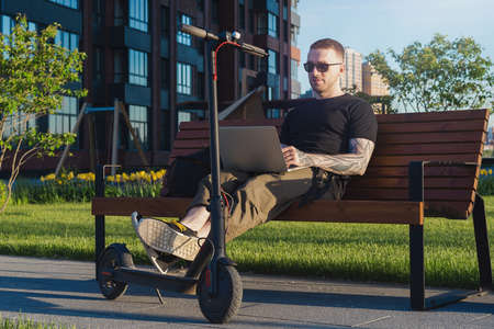 Young attractive man with laptop and modern electric kick scooter sitting at contemporary park at bench. and working Freelancer or business man working on laptop. Beautiful Sunset time.の写真素材