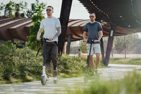Two men riding white and black electric kick scooters at beautiful park landscape. Men is on foreground, modern building and park is on background.の写真素材