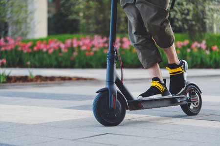 Close up of man riding black electric kick scooter at beautiful park landscape. Man is on foreground, modern building and park is on background.の写真素材