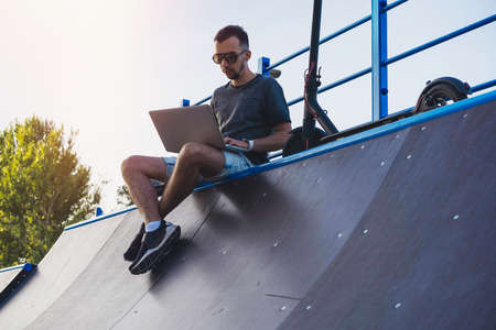 Young attractive man with laptop and modern electric kick scooter sitting at skater ramp in contemporary park. and working Freelancer or business man working on laptop. Beautiful Sunset time.の写真素材