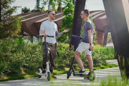 Two men talking next to white and black electric kick scooters at beautiful park landscape. Men is on foreground, modern building and park is on background.の写真素材