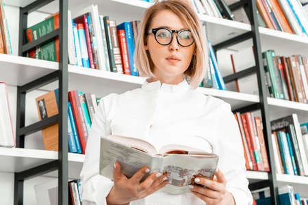 Young pensive attractive woman with glasses in formal clothes reading a book at library.の写真素材