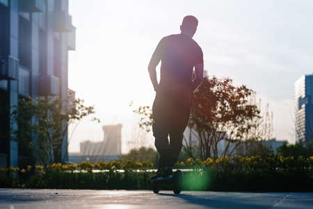 Man riding black electric kick scooter at beautiful cityscape at sunset. Man is on foreground, modern building, business or commercial centres are on background.の写真素材