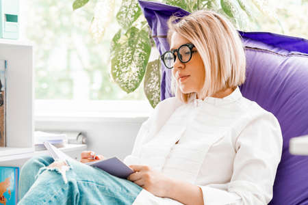 Young attractive smiling woman makes some notes in notepad at library and sitting on violet couch.の写真素材