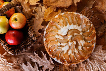 Freshly baked homemade apple pie near red apples in straw basket on yellow autumn leaves background. Autumn traditional pie, dish.の写真素材