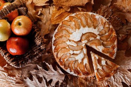 Freshly baked homemade apple pie near red apples in straw basket on yellow autumn leaves background. Woman hand holding piece of pie above plate, Rustic style. Top viewの写真素材