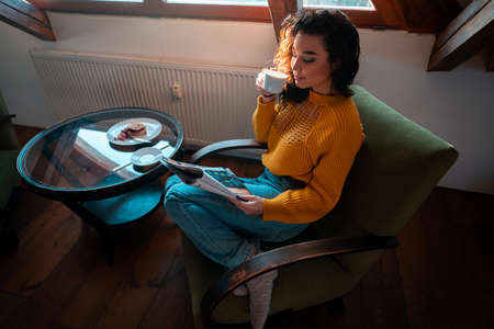 Young Attractive Woman in Yellow Cozy Sweater Sitting on Green Armchair, Drinking Hot Tea and...
