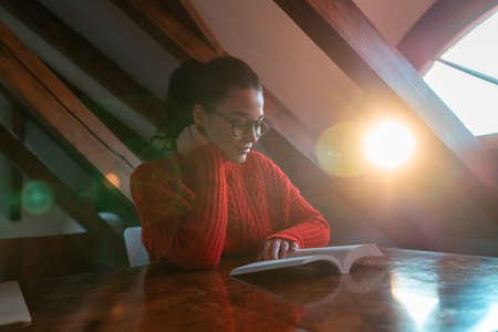 Beautiful young woman dressed in red cozy sweater reading book at attic mansard filled with sun light and smilingの写真素材