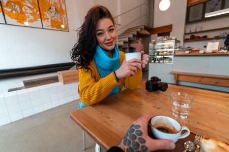 Young Woman in Yellow Sweater And Blue Scarf Drinking Hot Tea at Modern Cafe And Looking at Her Boyfriend. First Person View.の写真素材