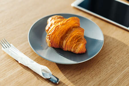 Close up of Freshly Baked Croissant on Blue Dish in Cafe and Tablet Near Utensils on Table. Concept...