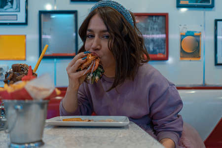 Hungry Attractive Woman Appetizingly Eating Burger At American Cafe. Enjoying Cheat Meal. American Fast Foodの写真素材