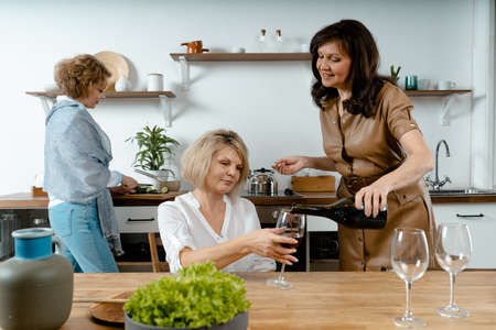 Beautiful Brunette Senior Lady Pouring Wine At Glass Of Her Old Friend at Modern Kitchen. Women Met For Preparing Meal. Living Full Life At Any Ageの写真素材