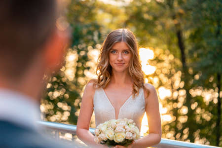 Smiling Happy Bride Holding Wedding Bouquet In Front Of Groom at Wedding Ceremony Outdoors At Park. Groom Is On Foreground And Blurred. Bride is On Background And on Focusの写真素材