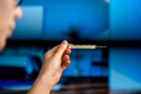 Woman Checking Body Temperature Using Mercury Thermometer In Blue Modern Interior. Health Research...