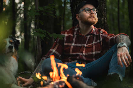 Bearded Hipster Man Near Bonfire In Dark Autumnal Forest an Looking At Sitting Sun. Travelling At Native Coutry, Vacation With Closed Bordersの写真素材