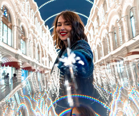 Beautiful Brunette Woman Holding Sparkler And Smiling Near Decorated With Christmas Lights Building. New Year And Christmas Celebration 2021の写真素材