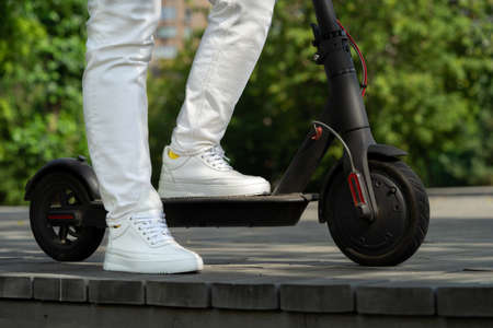 Close up of Young Woman In White Jeans And Sneakers Riding Black Electric Scooter On Wooden Plank Road At Park.の写真素材