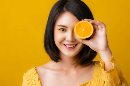 Beautiful portrait young Asian woman with orange fruit in studio yellow background. Isolated Asian girl smiling with holding orange fruit.の写真素材