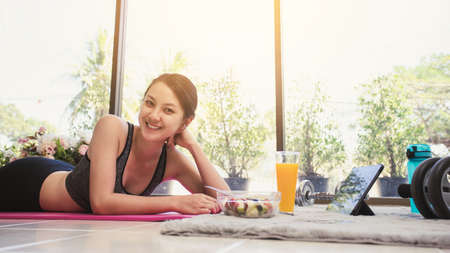 Healthy Asian woman smiling in sportswear lying on the floor and eating salad fruit at home. Eating diet and exercise relaxation concept.の写真素材