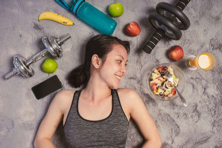 Healthy Asian woman smiling in sportswear lying on the floor with exercise equipment and salad fruit. Eating diet and exercise relaxation concept.の写真素材