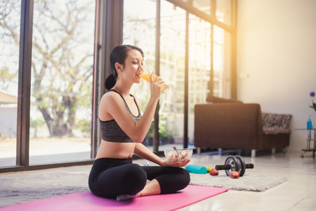 Healthy Asian woman in sportswear sitting on the yoga mat drinking orange juice and eating salad fruit at home. Eating diet and exercise relaxation concept.の写真素材