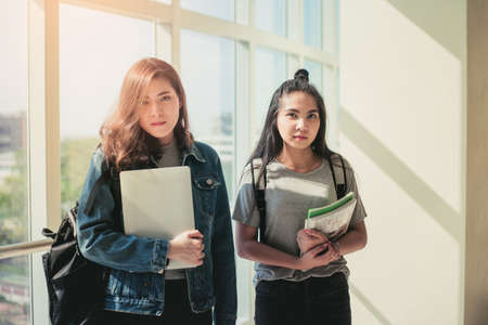 Asian young woman student talking with friend in library and holding books on her arms. education concept.の写真素材