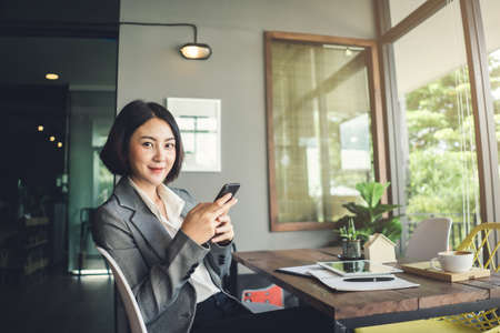 Asian businesswoman smile working and holding smartphone sitting in coffee shop at wooden table.の写真素材