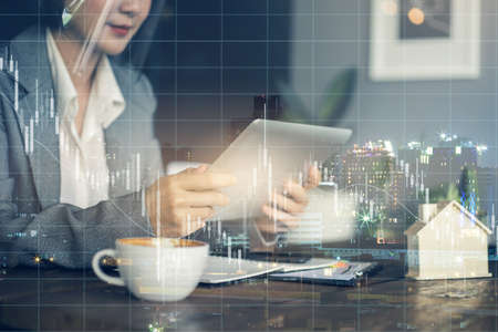 Double exposure hand of business asian woman holding use tablet with trend graph and cityscape. Real estate business impact from the crisis.の写真素材