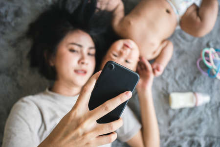 Closeup hand holding mobile phone for selfie, New normal, Happy Asian mom and baby newborn lying and use smartphone for social distance stay at home.の写真素材