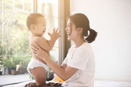 Asian mom smiling happy holding and playing with her son in living room. Mom and baby clap your hands together. Mom and baby in motherâs day concept.の写真素材