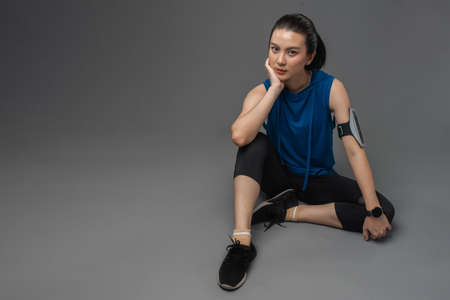 Beautiful young Asian woman in sportswear after exercise sitting and resting on the floor looking camera on grey background.の写真素材