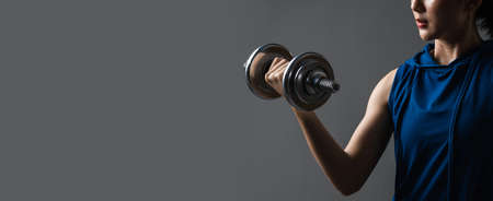 Selective focus steel dumbbell on hand of Asian woman workout with lifting weight on web banner grey background. Young athletic woman pumping up muscules with dumbbells.の写真素材