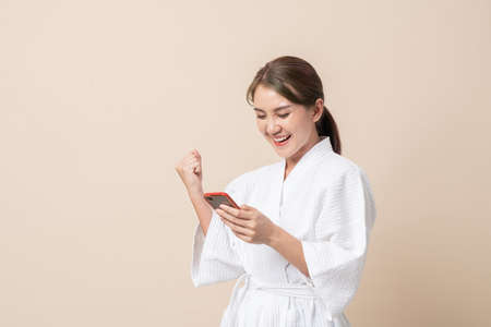 Portrait Beautiful young Asian women happy smile in bathrobe or spa suit and using smartphone trading or chatting on brown isolated studio background.の写真素材