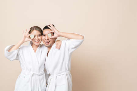 Happy Asian couple smile in bathrobe or spa suit with cucumber slice for facial mask together on brown isolated studio background.の写真素材