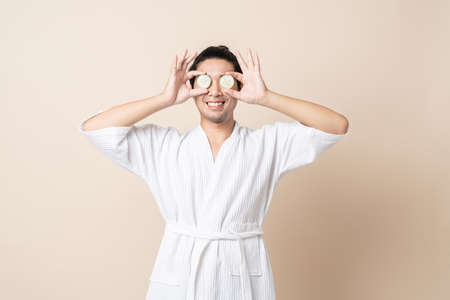 Portrait Asian man happy smile in bathrobe or spa suit with holding cucumber slice for facial face mask on brown isolated studio background.の写真素材