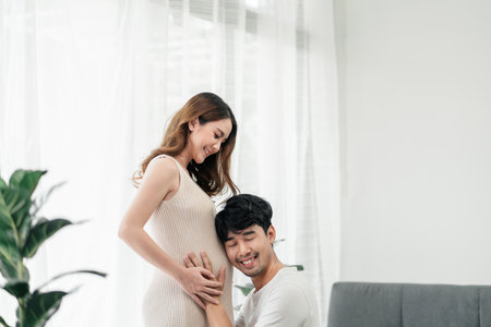 A happy pregnant asian couple. The husband is very happy while listening to his baby's sound in his wife's tummy in the living room.の写真素材