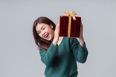 Happy beautiful Asian woman smiling in casual green sweater holding a gift box for anniversary or new year on isolated grey background. Happy anniversary or new year festival concept.の写真素材