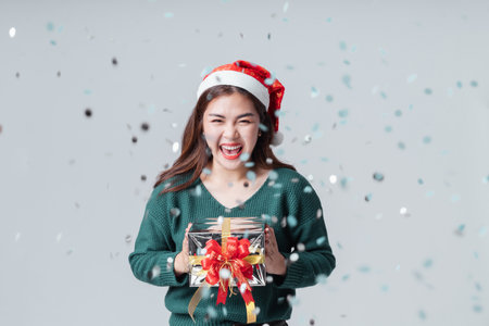 Celebration Cheerful Asian woman in casual green sweater and christmas santa hat holding a giftbox isolated grey background. Happy anniversary or new year festival concept.の写真素材