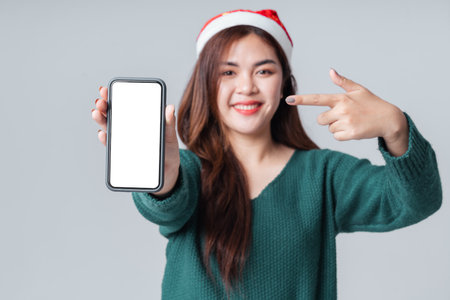 Selective focus cell phone blank screen, Asian woman in casual green sweater and Christmas Santa hat, showing smartphone empty space for mobile app on screen. Happy anniversary or new year concept.の写真素材