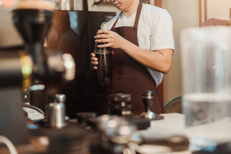 Barista Asian man holds in hands steel shaker at coffee shop. Asian man barista in white t-shirt and apron working at coffee shop. SME business coffee shop concept.の写真素材