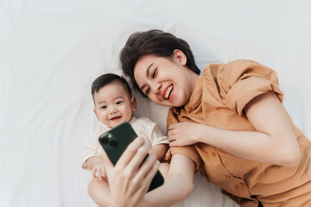 Happy Asian mother and her newborn baby making a selfie or video call to father or relatives in a bed. Concept of technology, new generation,family, connection, parenthoodの写真素材