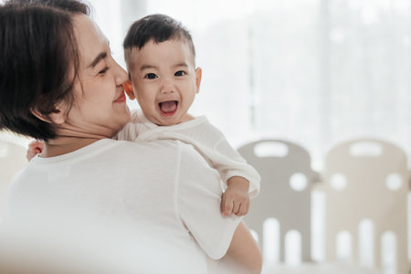Happiness of motherhood. Young Asian mom with adorable baby on his hands standing near window at home. Loving mother spending time with infant child. Mother's day concept.の写真素材