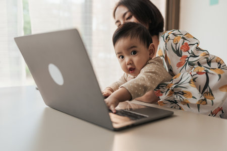 A little boy Asian baby is disturbing his mother while she is working at home. Asian mother carrying her son baby while working on computer laptop at home. Long distance working from home concept.の写真素材